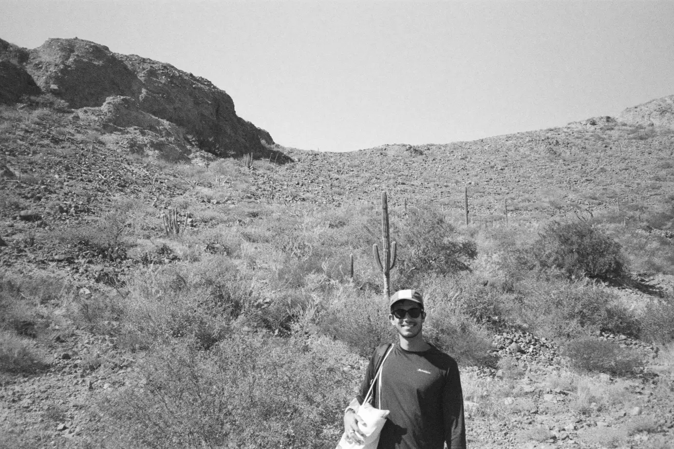 A black and white photo of me smiling in a cap and sunglasses, in front of a desert hillside with cacti